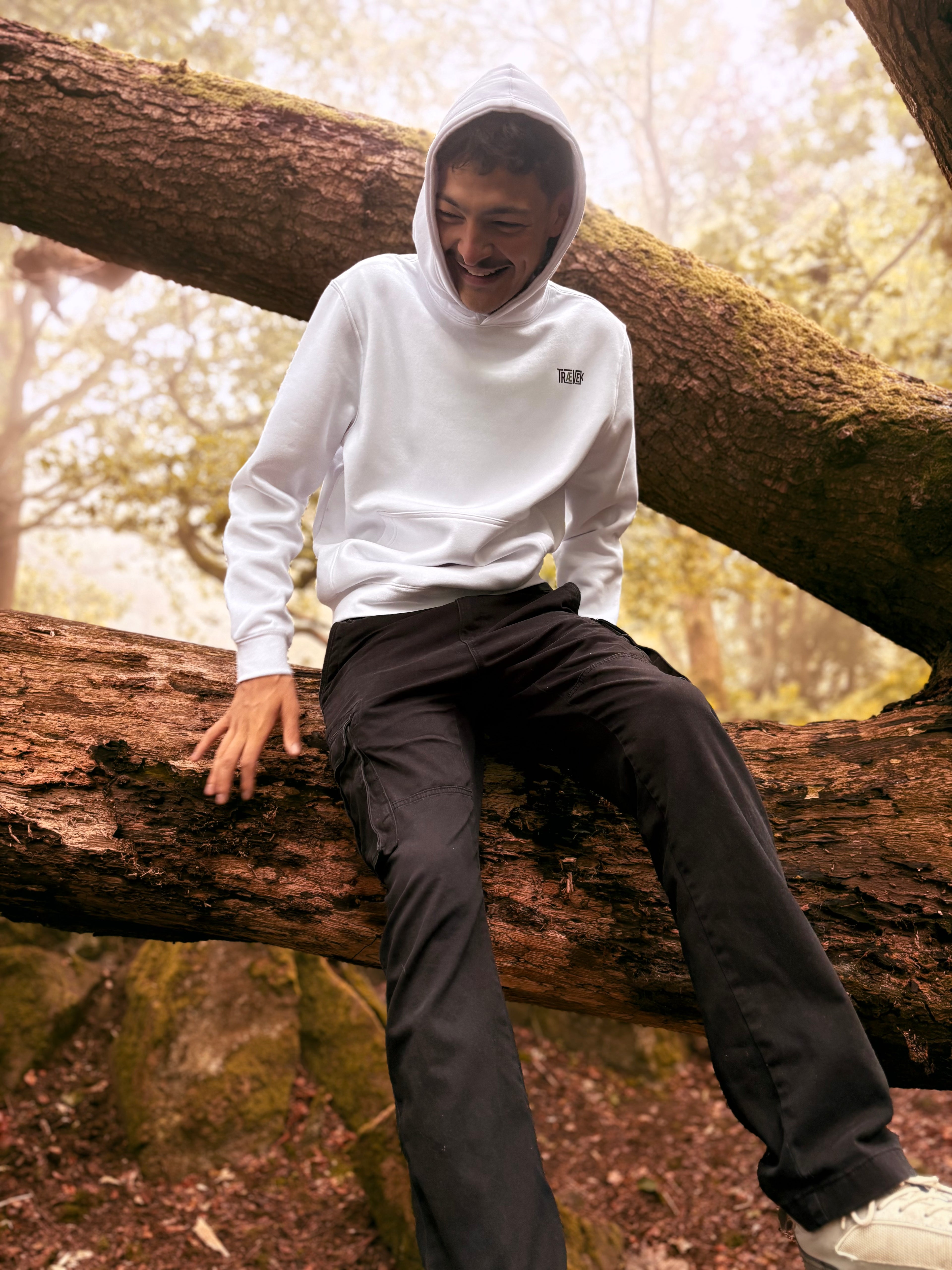 A man wearing a white hoodie with a small black 'TRAEVEK' logo on the chest and black pants, sitting on a large fallen tree trunk in a forest.
