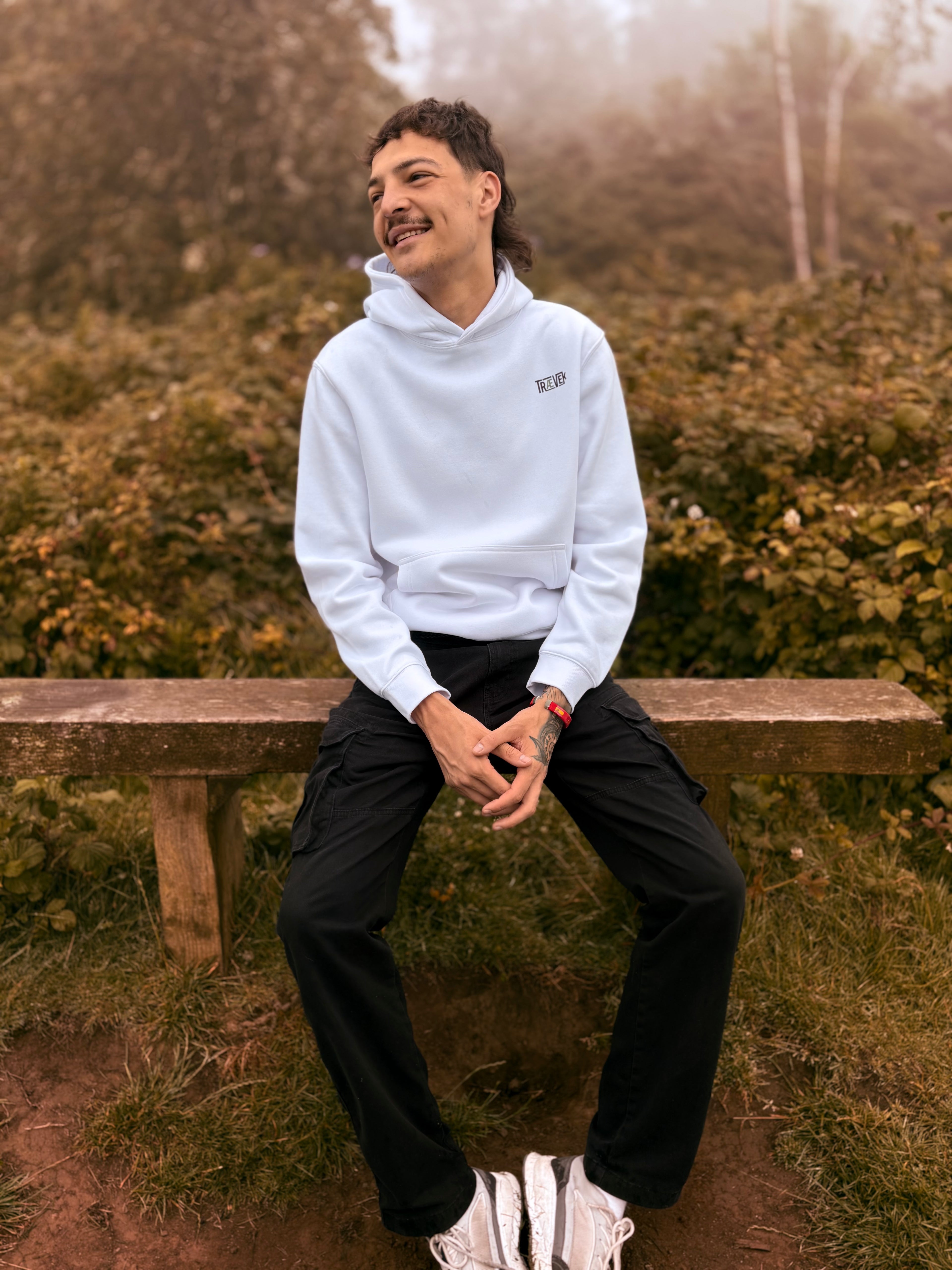 A man wearing a white pullover hoodie with a small black logo on the chest, black cargo pants, and white sneakers sits on a wooden bench in an outdoor, misty forest setting.